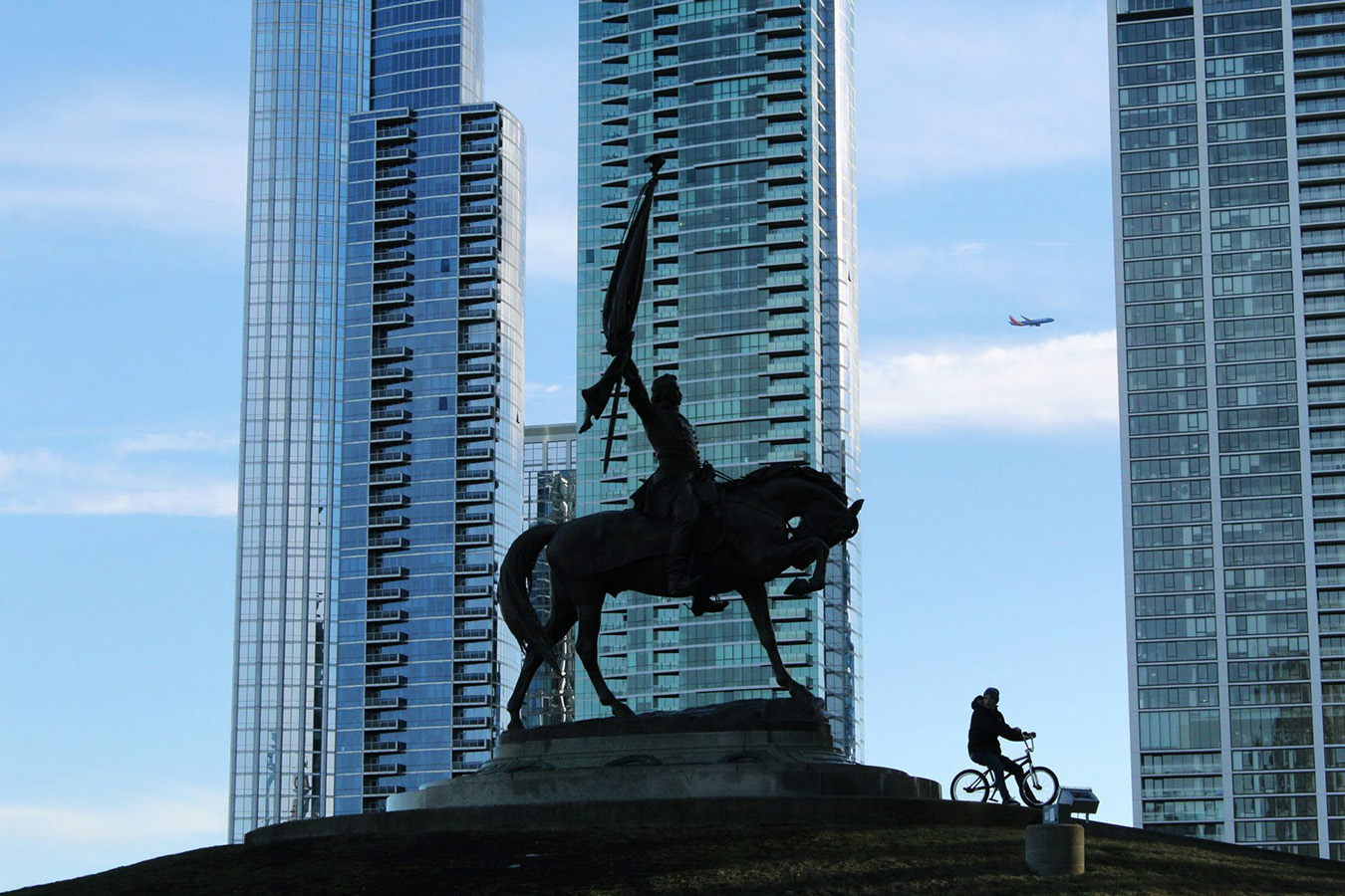 photo of statue of person on horseback a real person on a bicycle and an airplane overhead