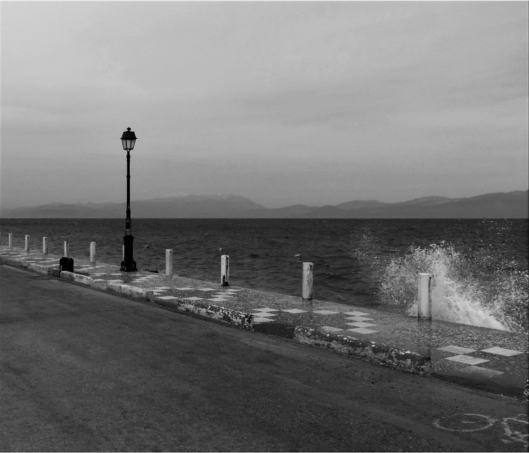 photograph of a road and lamp post at edge of water with one small wave splashing up
        			 over the edge of the tiled walkway